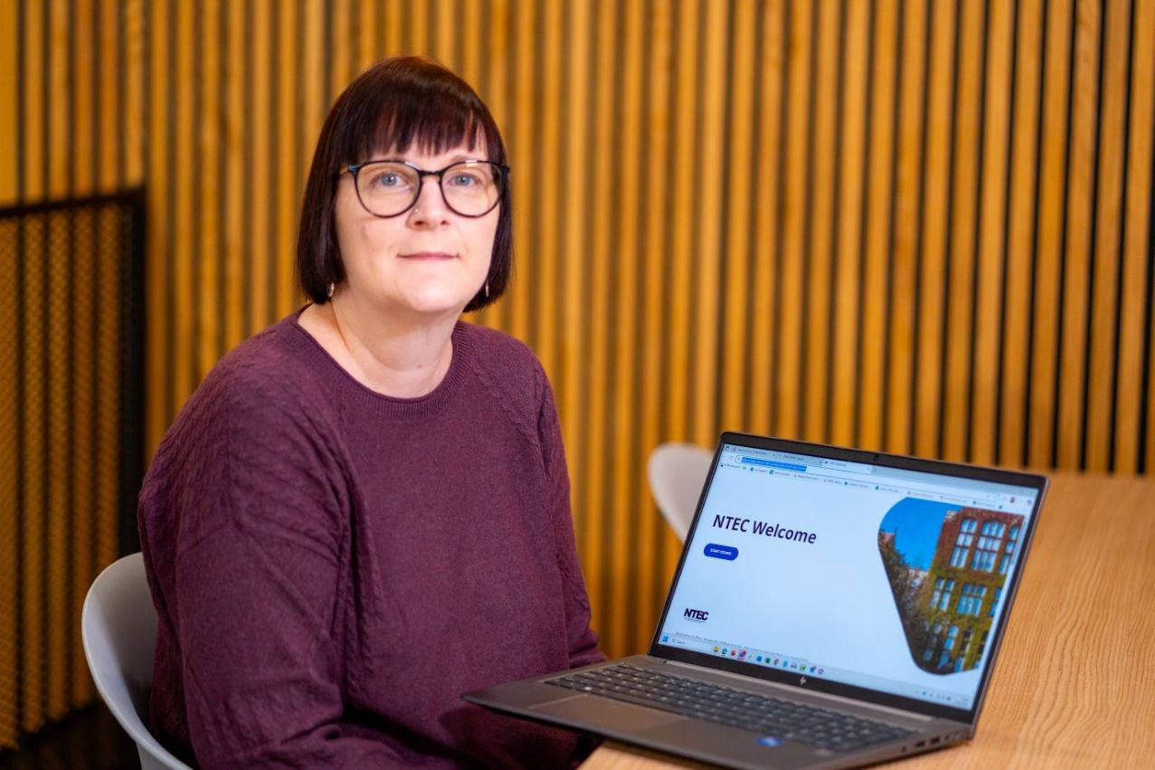 Image of Caroline Hughes, who is sitting looking at the camera with her laptop. Caroline has short black hair wearing glasses and a burgundy jumper.